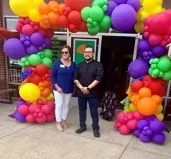 A man and a woman are standing in front of a rainbow colored balloon arch.