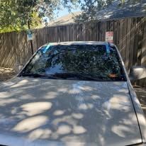 Silver car parked in front of a wooden fence. The windshield has blue tape on it.