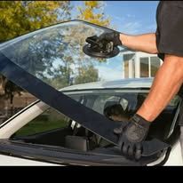A person in gloves using a suction cup to remove a car windshield. The car is outside in a sunny setting.