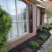 Exterior of a brick house with large windows, black shutters, and a small garden with mulch and green plants.