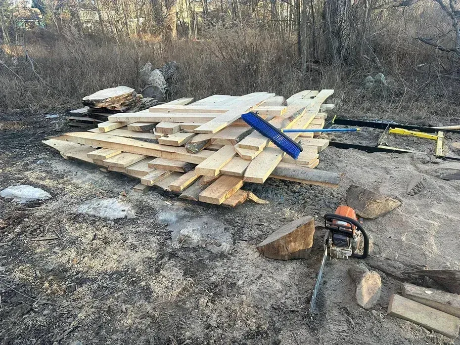 A stack of lumber rests on the ground in a clearing, with a chainsaw and blue brush lying on top of the boards.