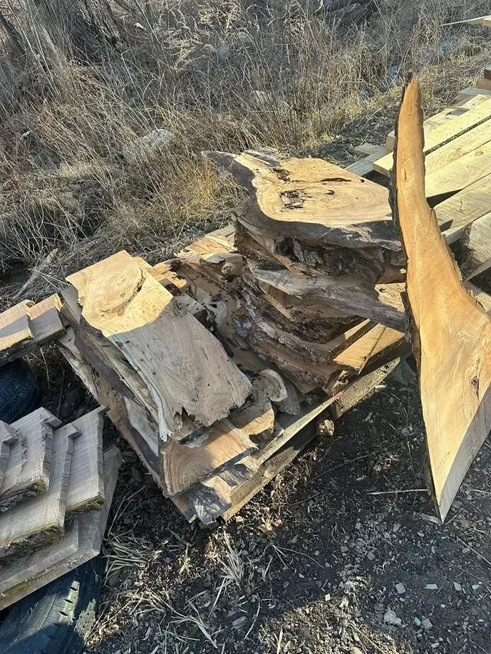 A stack of rough-cut wooden slabs and boards sitting on a pallet outdoors on dark, dirt-covered ground.