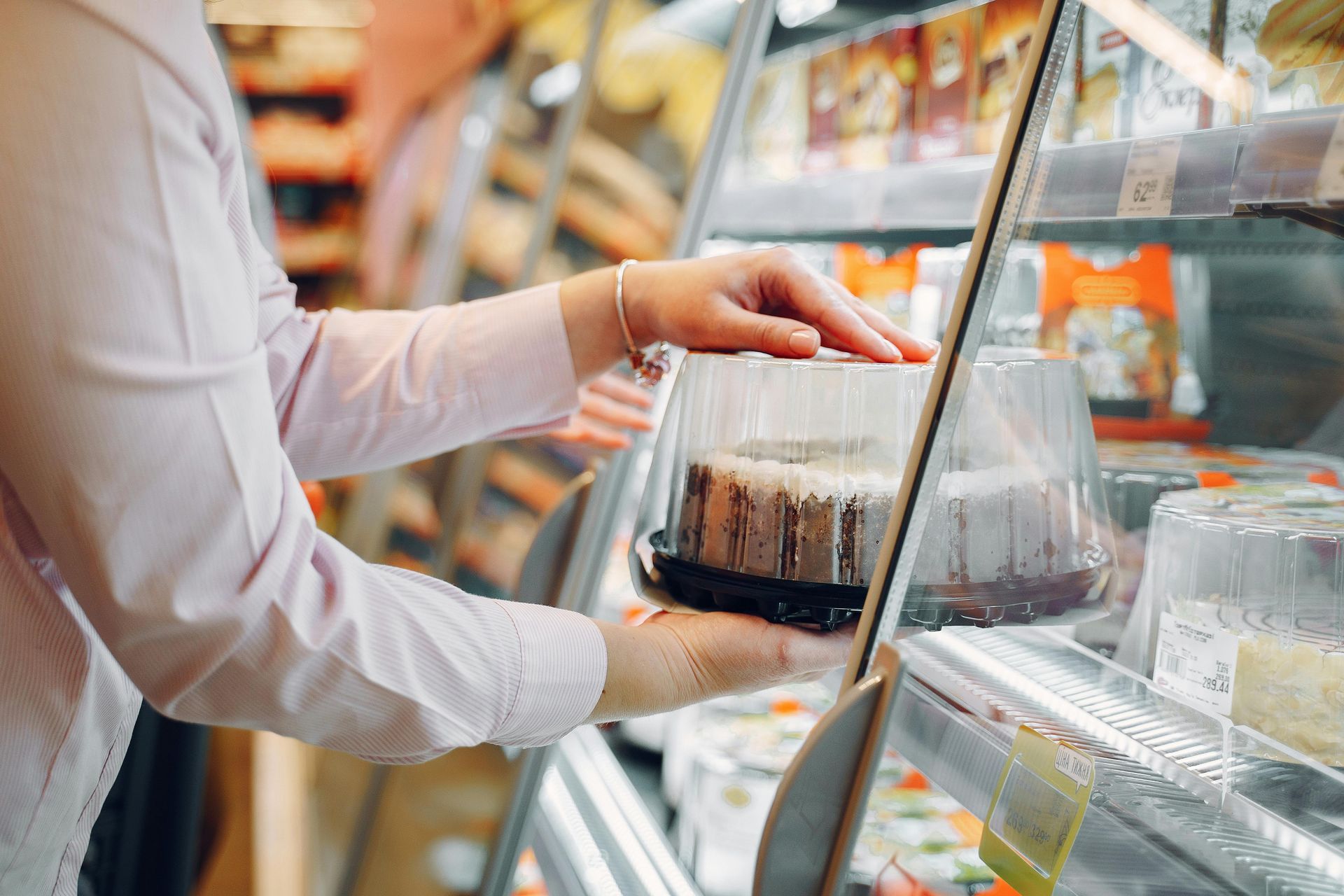 Person in pink shirt selecting a cake from a refrigerated display case in a grocery store.