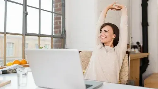 A woman is stretching her arms while sitting at a desk in front of a laptop computer.