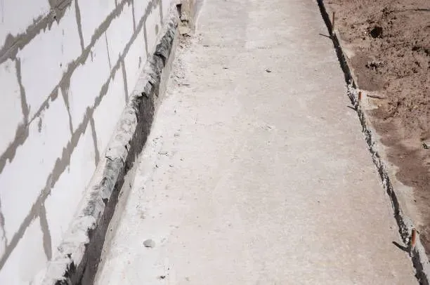 Exterior view of a narrow concrete drainage trench or French drain installed alongside a white brick foundation wall.
