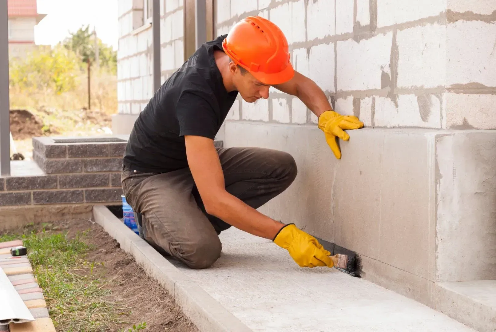 Contractor in safety gear applying waterproof sealant to the base of a concrete slab foundation.