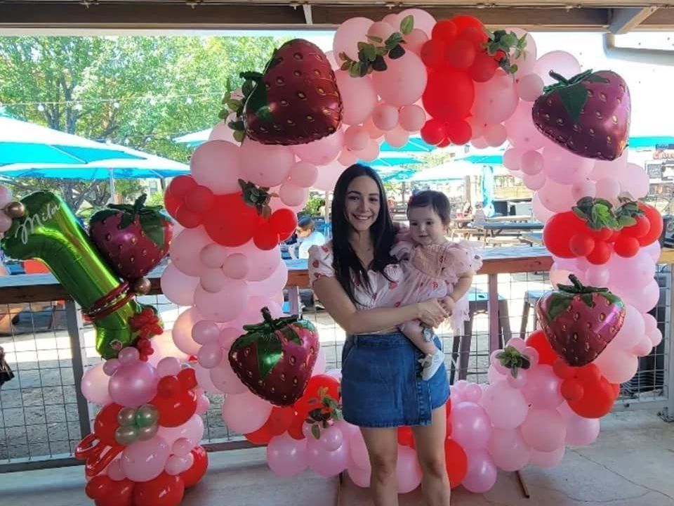 A woman is holding a baby in front of a strawberry balloon arch.