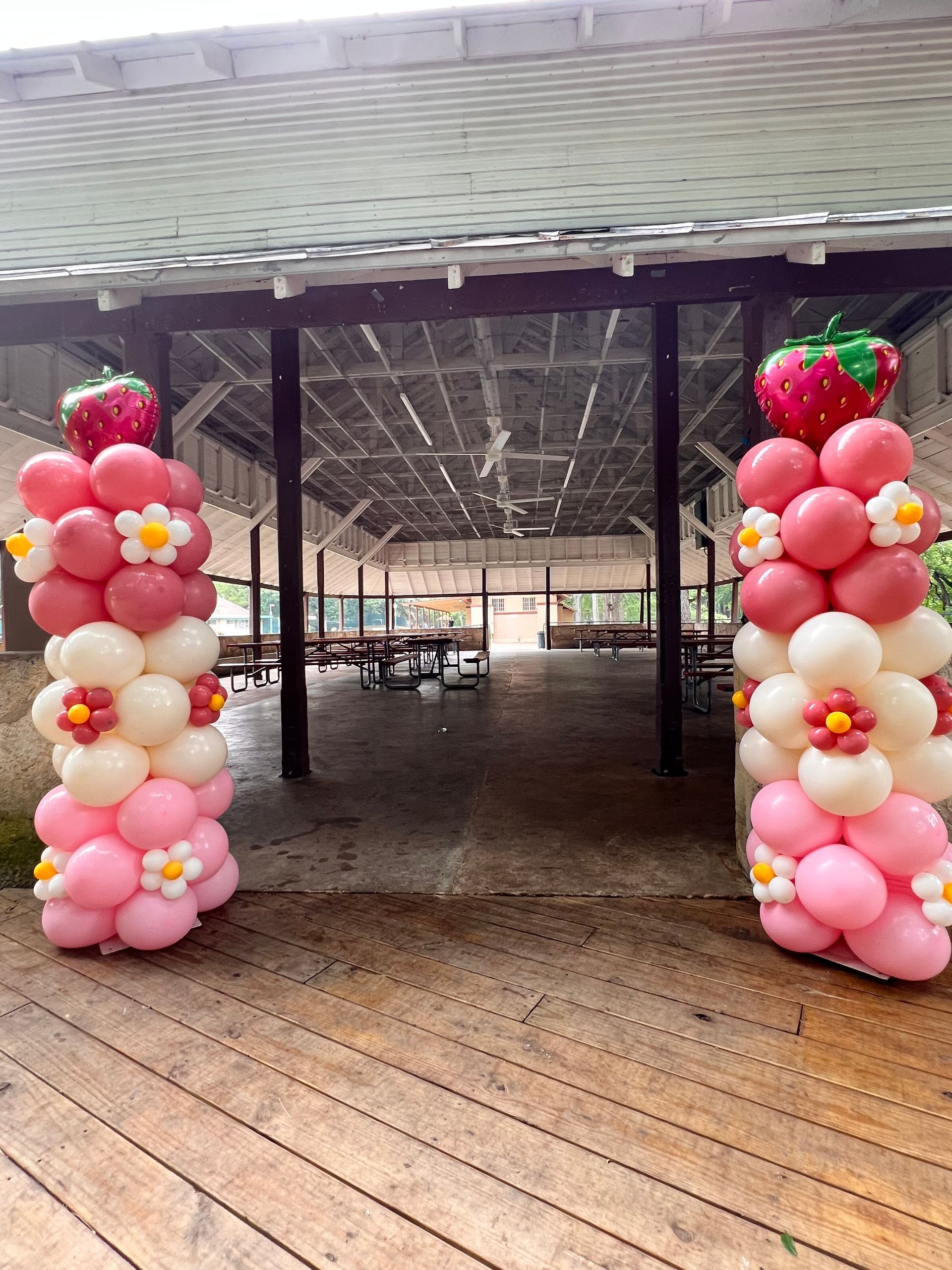 Two columns of pink and white balloons with strawberries and flowers are sitting on a wooden deck.