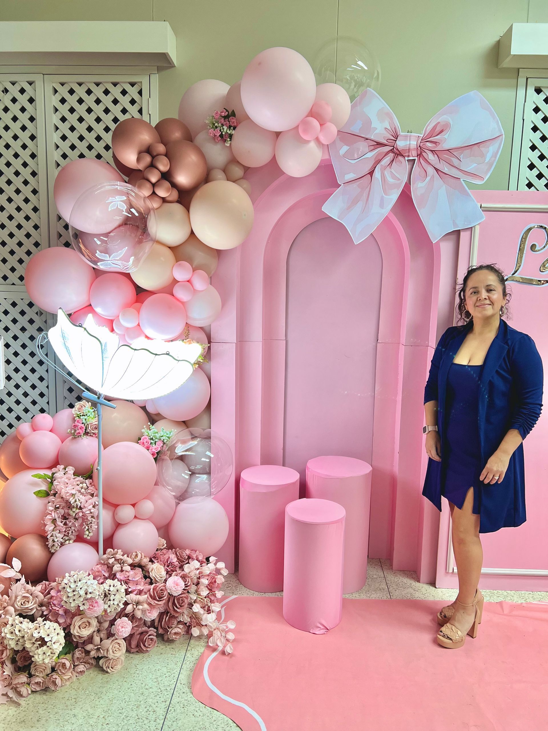 A woman is standing in front of a pink wall filled with pink balloons and flowers.