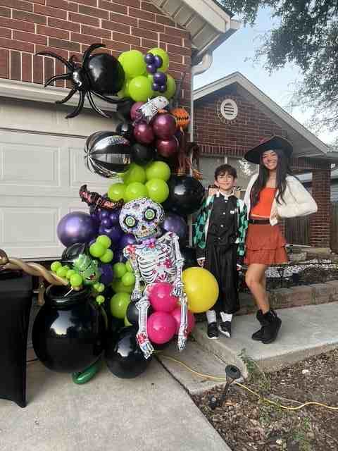 Two kids in costume pose next to a Halloween balloon archway with a spider and skeleton in front of a brick house.