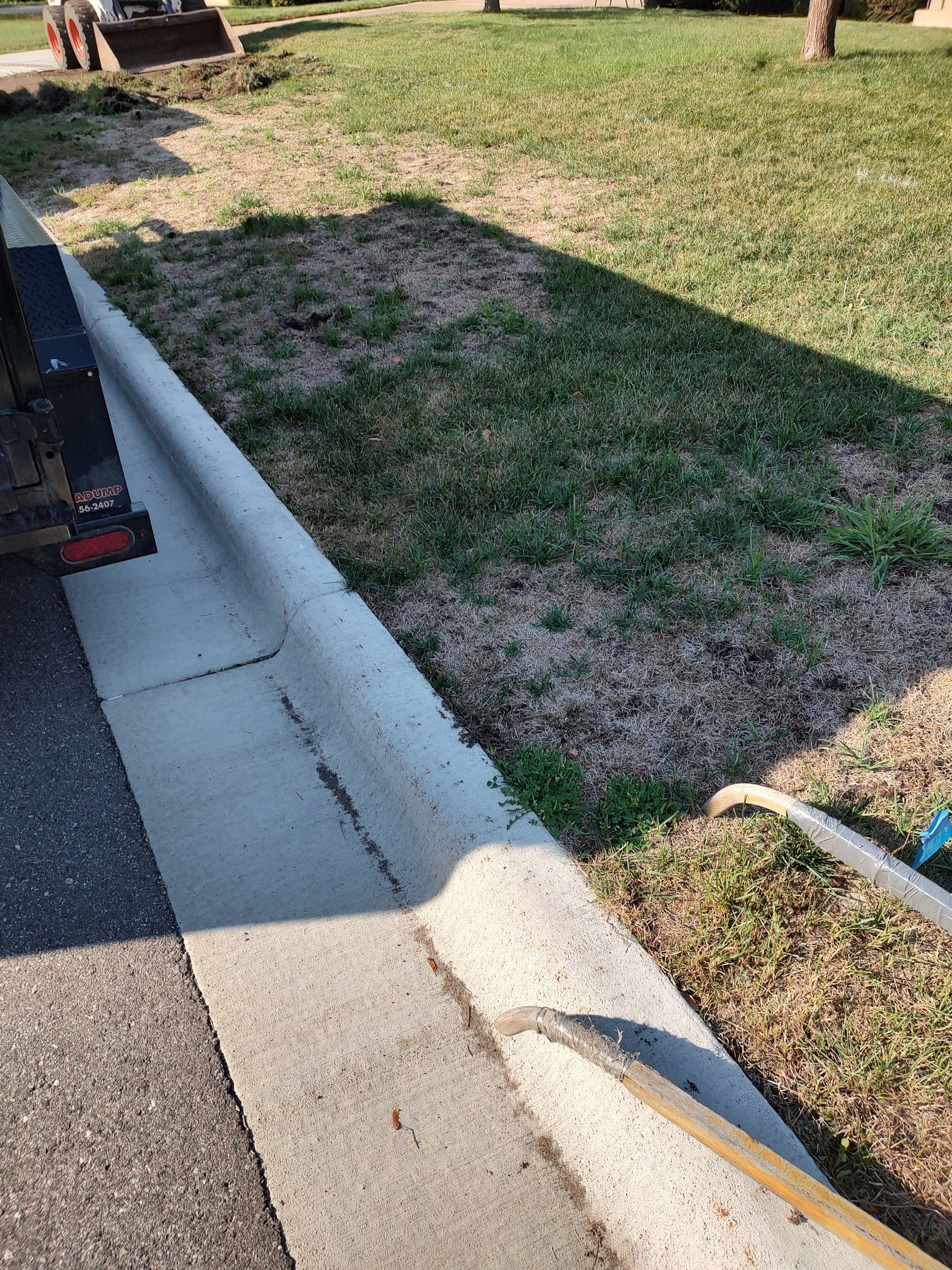 Concrete curb next to grass with a hose lying on the ground. Trailer on the left.