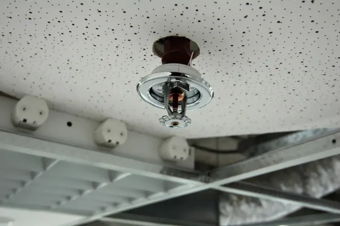 Testa di un impianto antincendio montata su un soffitto bianco screziato. Sotto, travi strutturali e apparecchi elettrici a vista.