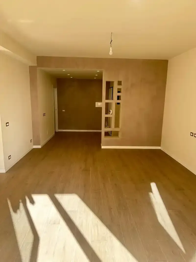 Interno vuoto di una stanza con pavimenti in legno marrone e pareti beige; la luce del sole proietta ombre.