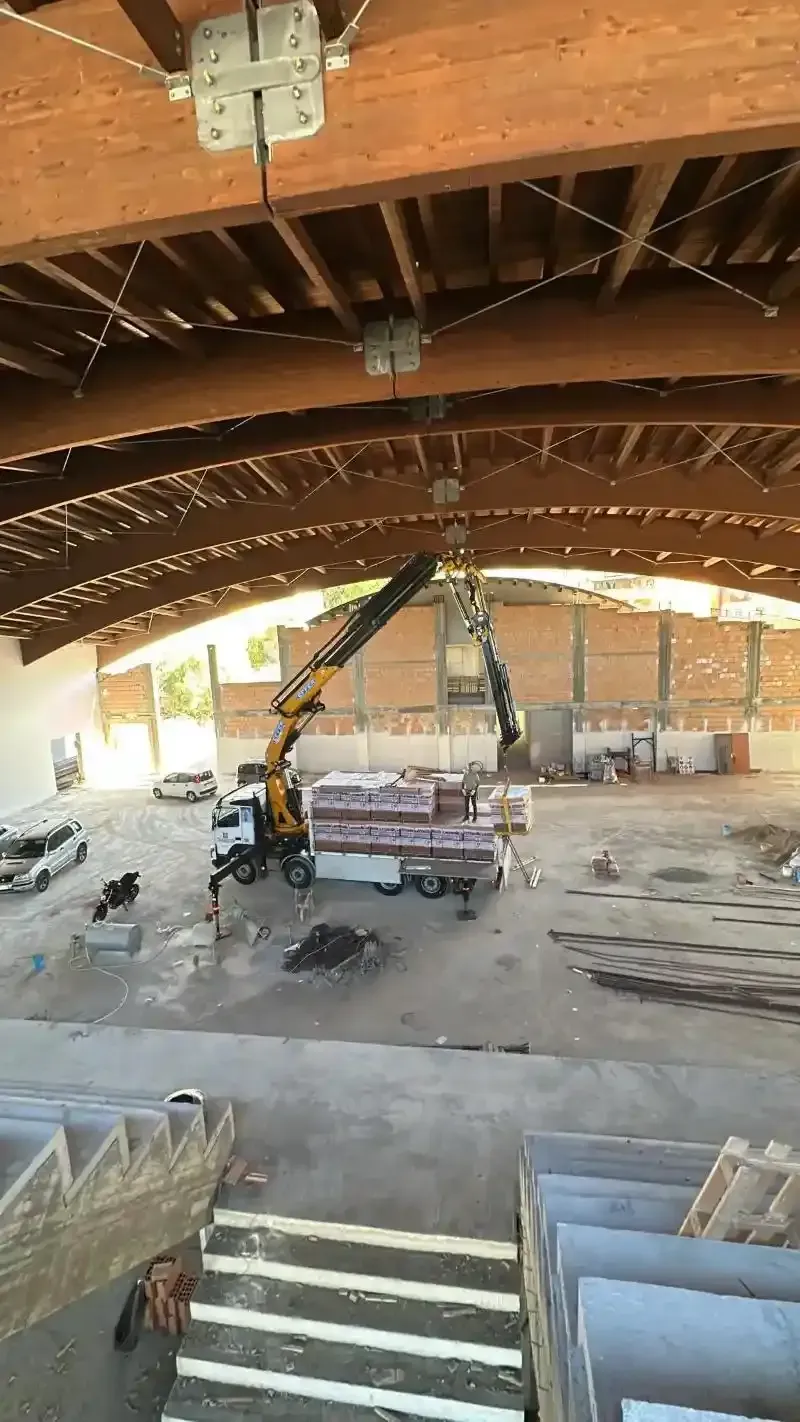 Una gru solleva materiali da costruzione all'interno di un capannone incompiuto con travi di legno.