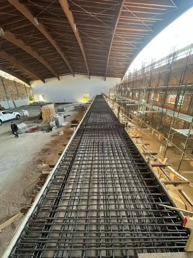 Cantiere con struttura in tondino di ferro, sotto una struttura di copertura curva. Impalcature sulla destra, operai e veicoli sullo sfondo.