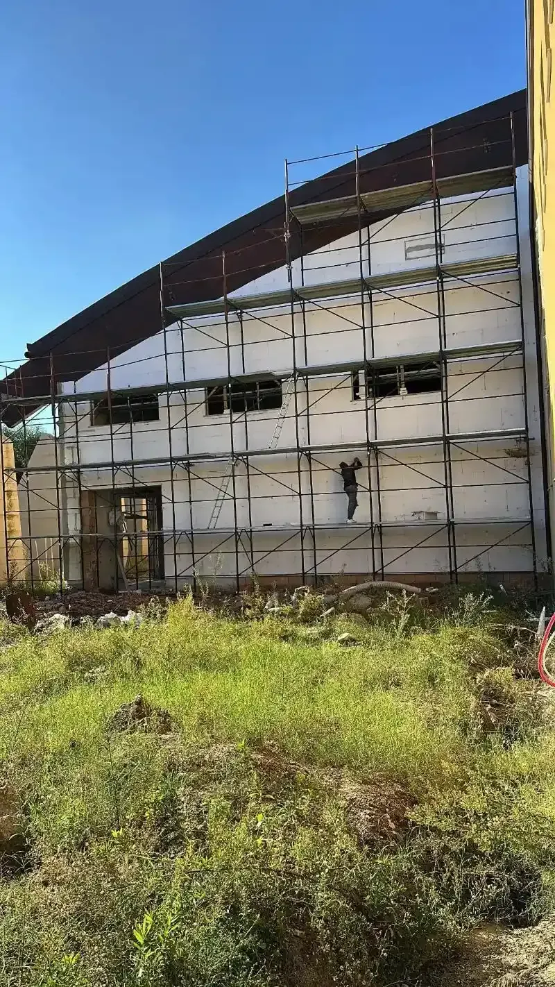 Esterno di un edificio con impalcature; lavori di costruzione. Erba verde in primo piano, cielo azzurro sullo sfondo.