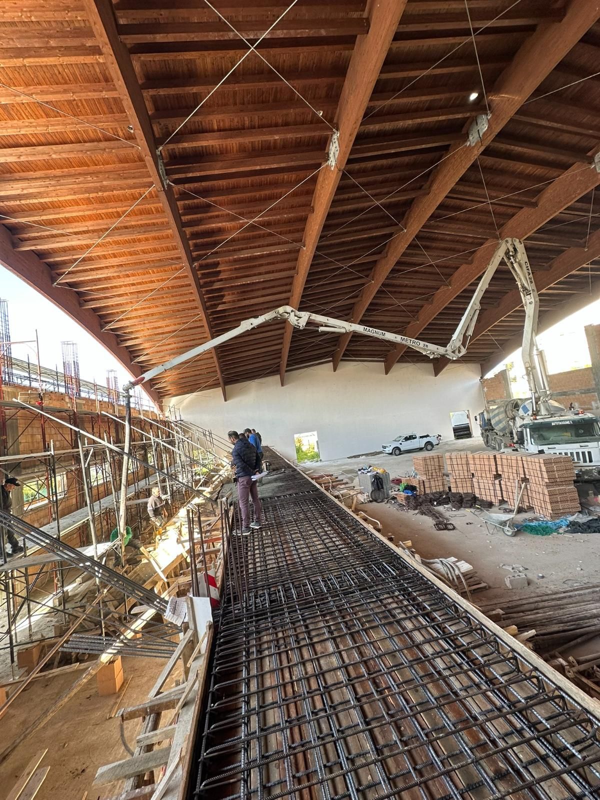 Cantiere: operai vicino alle barre d'armatura, pompa per calcestruzzo che versa il calcestruzzo sulla struttura, sotto un tetto in legno.