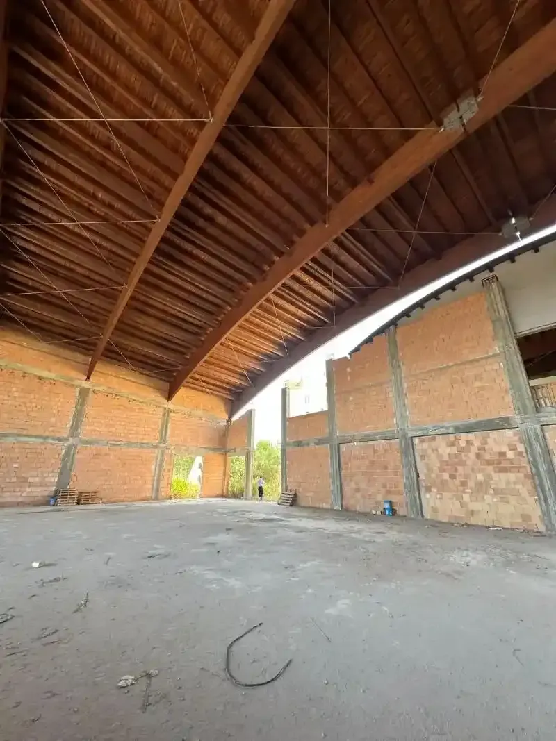Vista interna di un edificio incompiuto con pareti in mattoni a vista, tetto ad arco in legno e pavimento in cemento.