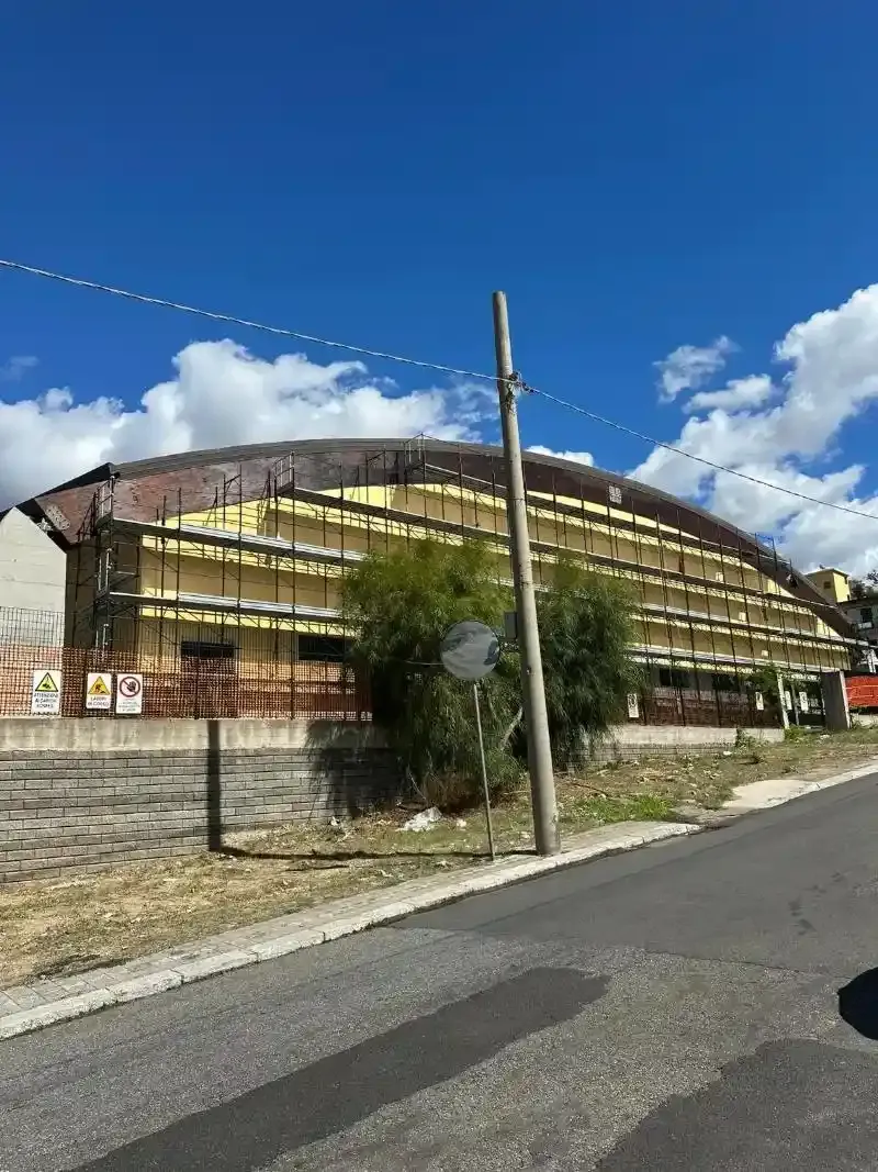 Edificio giallo con tetto curvo, dietro una recinzione lungo la strada, sotto un cielo azzurro con nuvole.