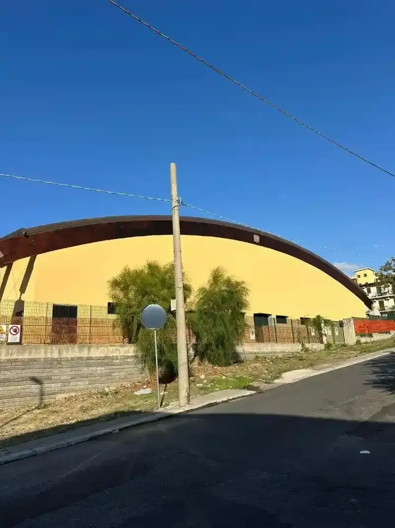 Edificio ad arco giallo, cielo azzurro, palo della luce in primo piano, strada, alberi.