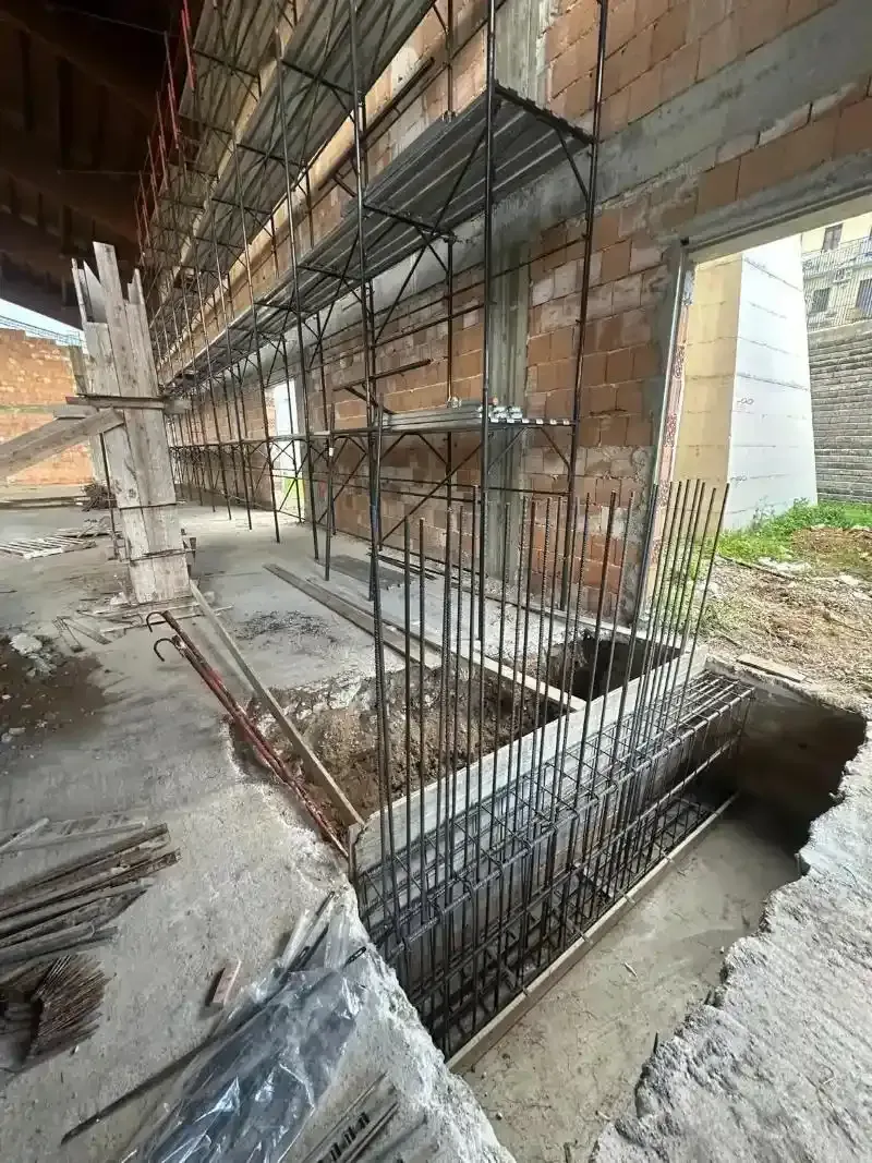 Cantiere con barre d'armatura a vista nelle fondamenta e nei ponteggi. Muri in mattoni, pavimento in cemento, ambiente esterno.