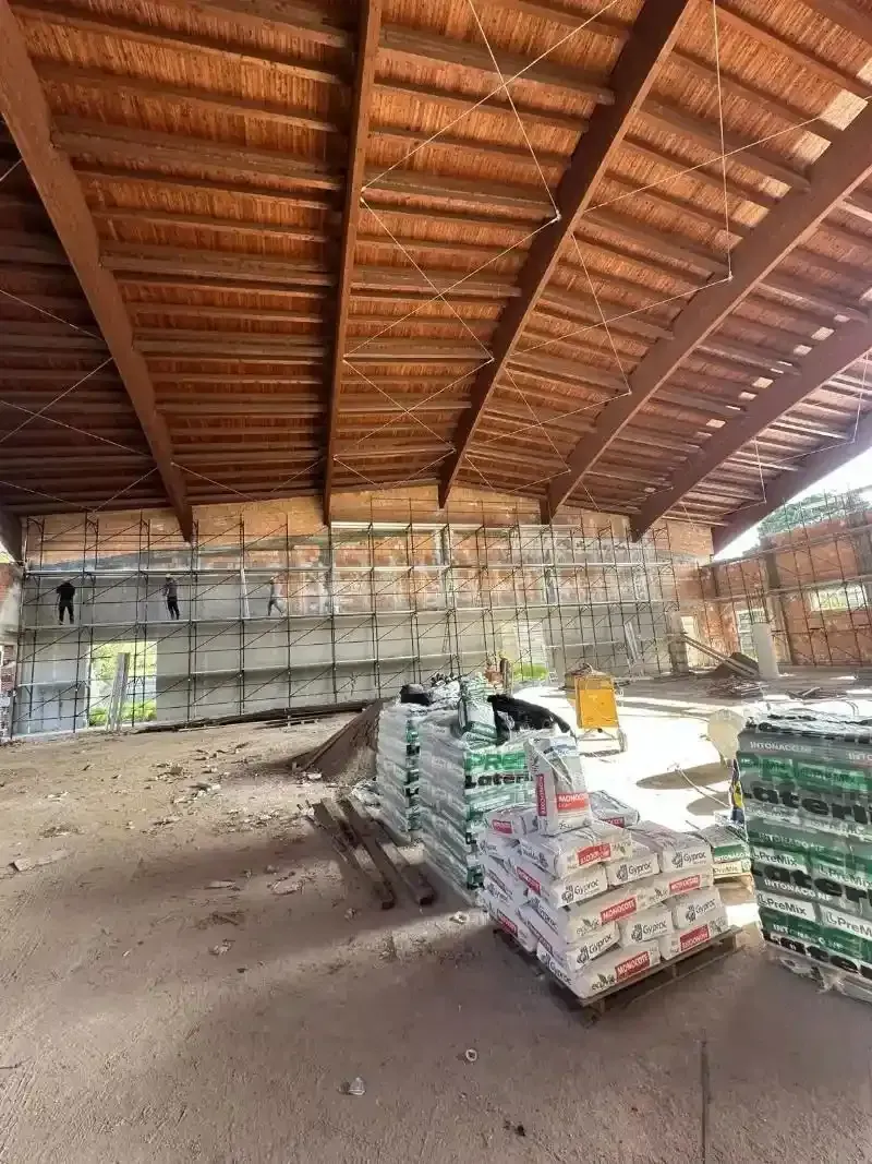 Interno di un edificio in costruzione, con soffitto in legno a vista e pile di materiali sul pavimento.