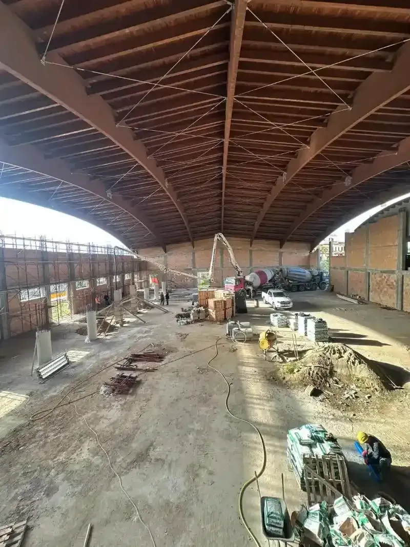 Cantiere sotto un grande tetto ad arco in acciaio. Sono presenti mattoni, attrezzature e operai.