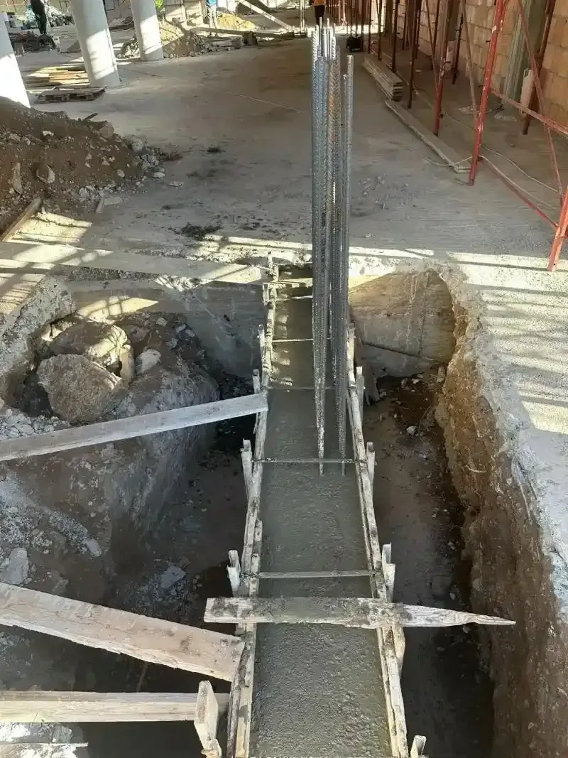 Cantiere con calcestruzzo gettato in una trincea, barre di armatura visibili. Casseforme in legno e impalcature sullo sfondo.