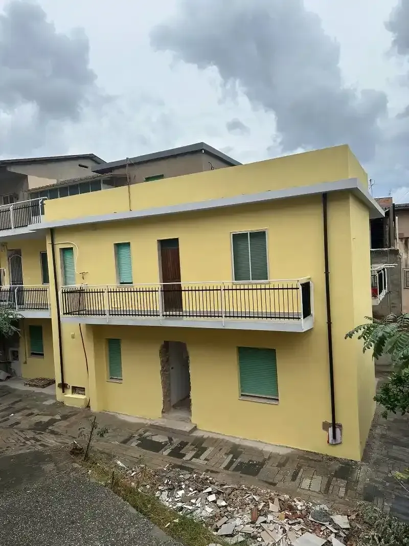 Edificio giallo a due piani con persiane verdi, un balcone e una porta marrone; cielo nuvoloso.