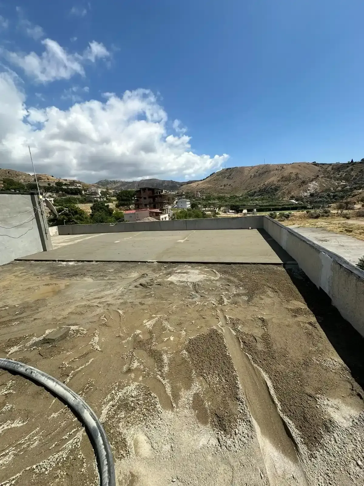 Tetto in cemento con vista sulla città e sulle montagne sotto un cielo azzurro con nuvole bianche.