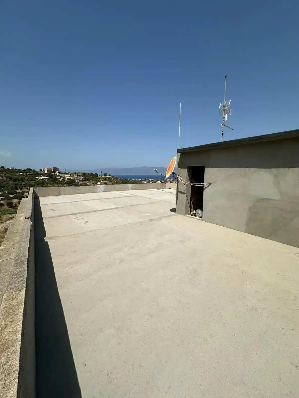 Tetto in cemento con porta, antenne e vista sul mare in lontananza sotto un cielo azzurro.
