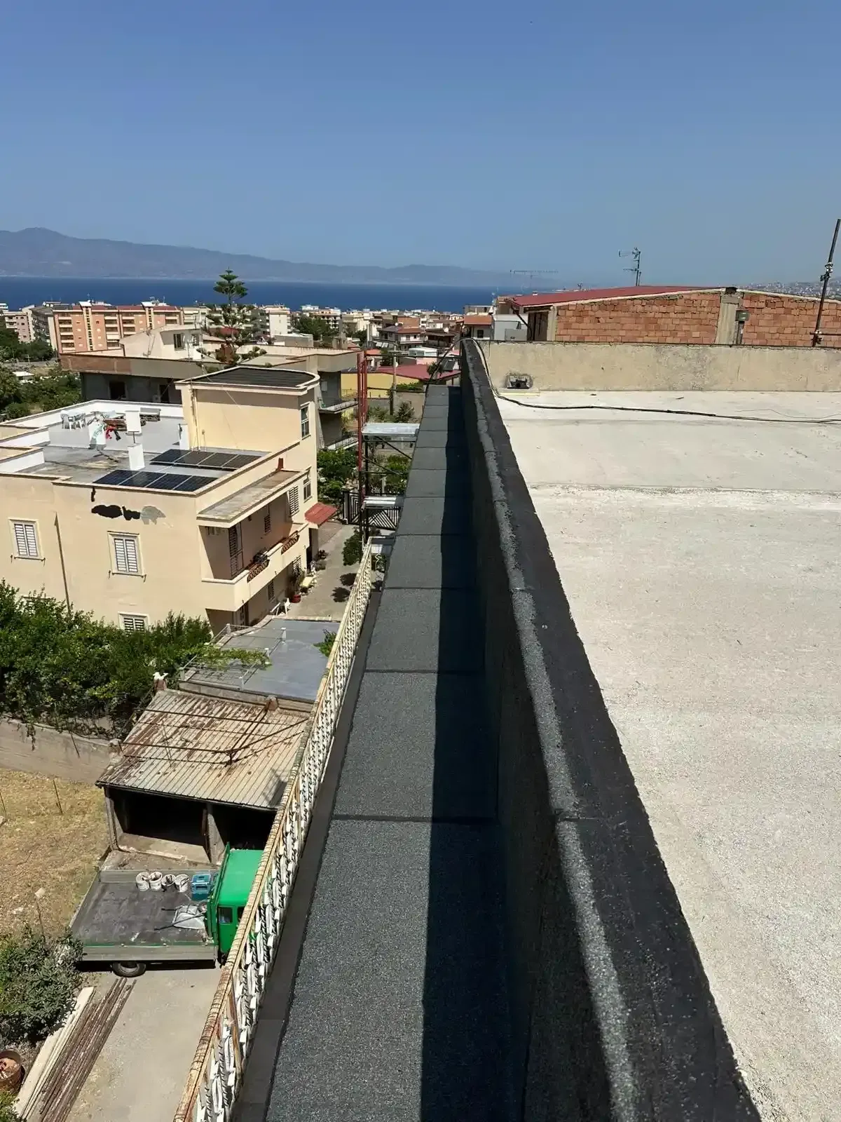 Vista da un tetto: un parapetto bordato di nero si estende fino all'orizzonte, dominando una città e il mare sotto un cielo azzurro.