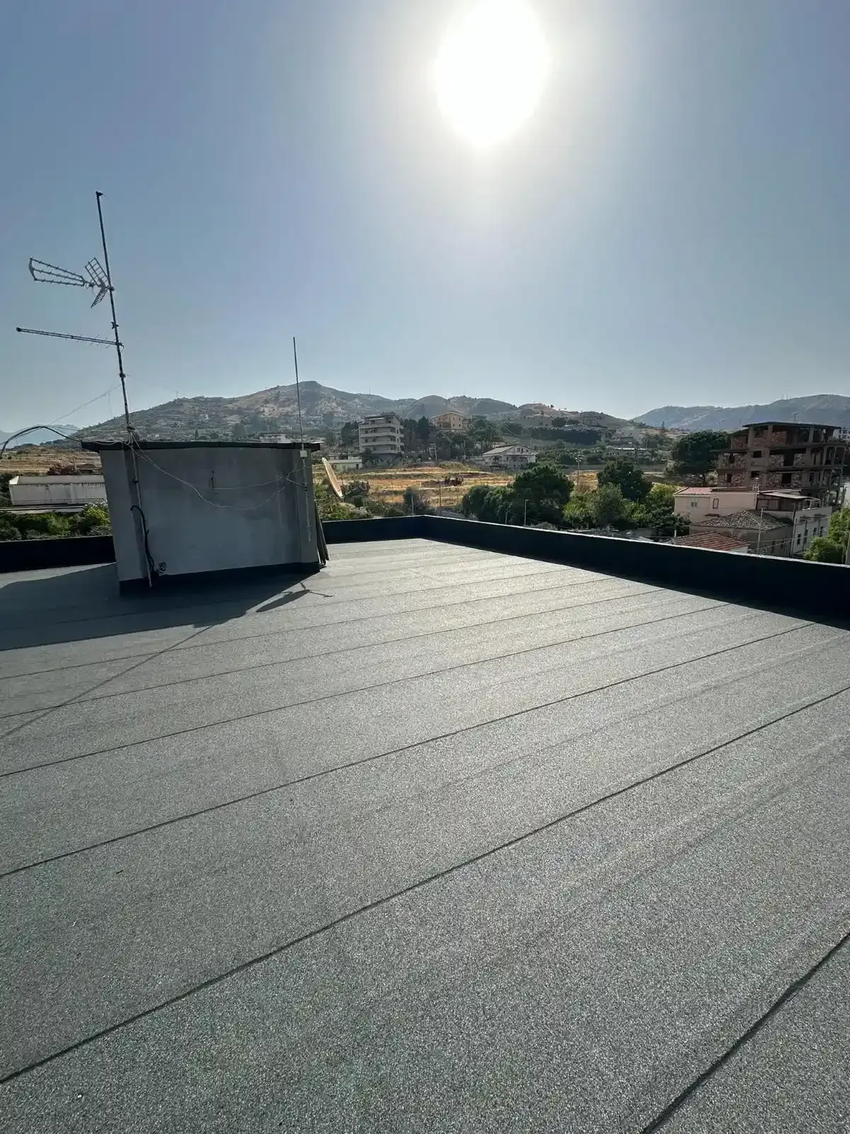 Un tetto piatto e nero con una piccola struttura metallica, un'antenna, montagne e il sole nel cielo.
