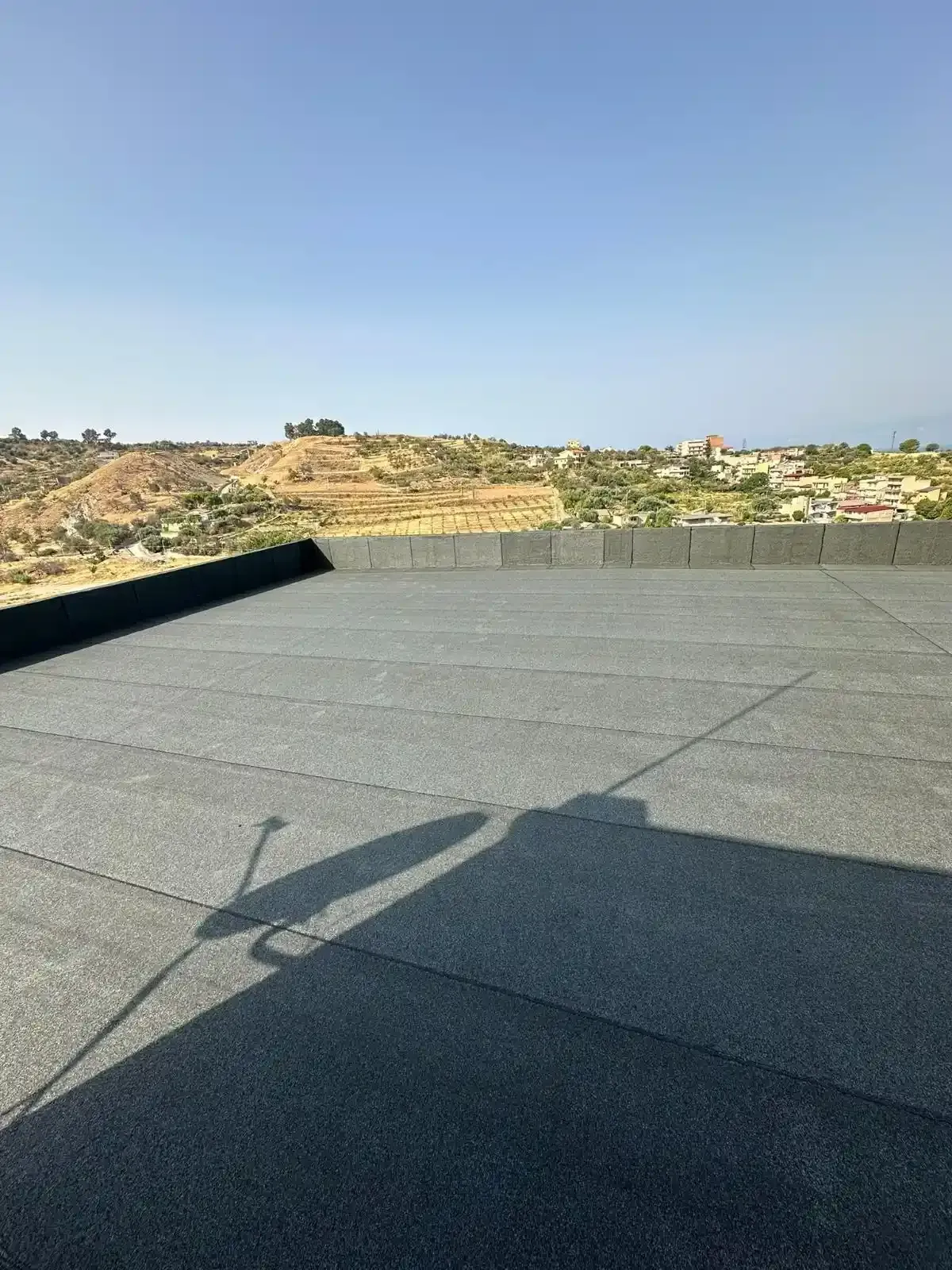 Un tetto piatto e grigio con un'antenna parabolica che proietta la sua ombra. Paesaggio collinare e cielo azzurro sullo sfondo.