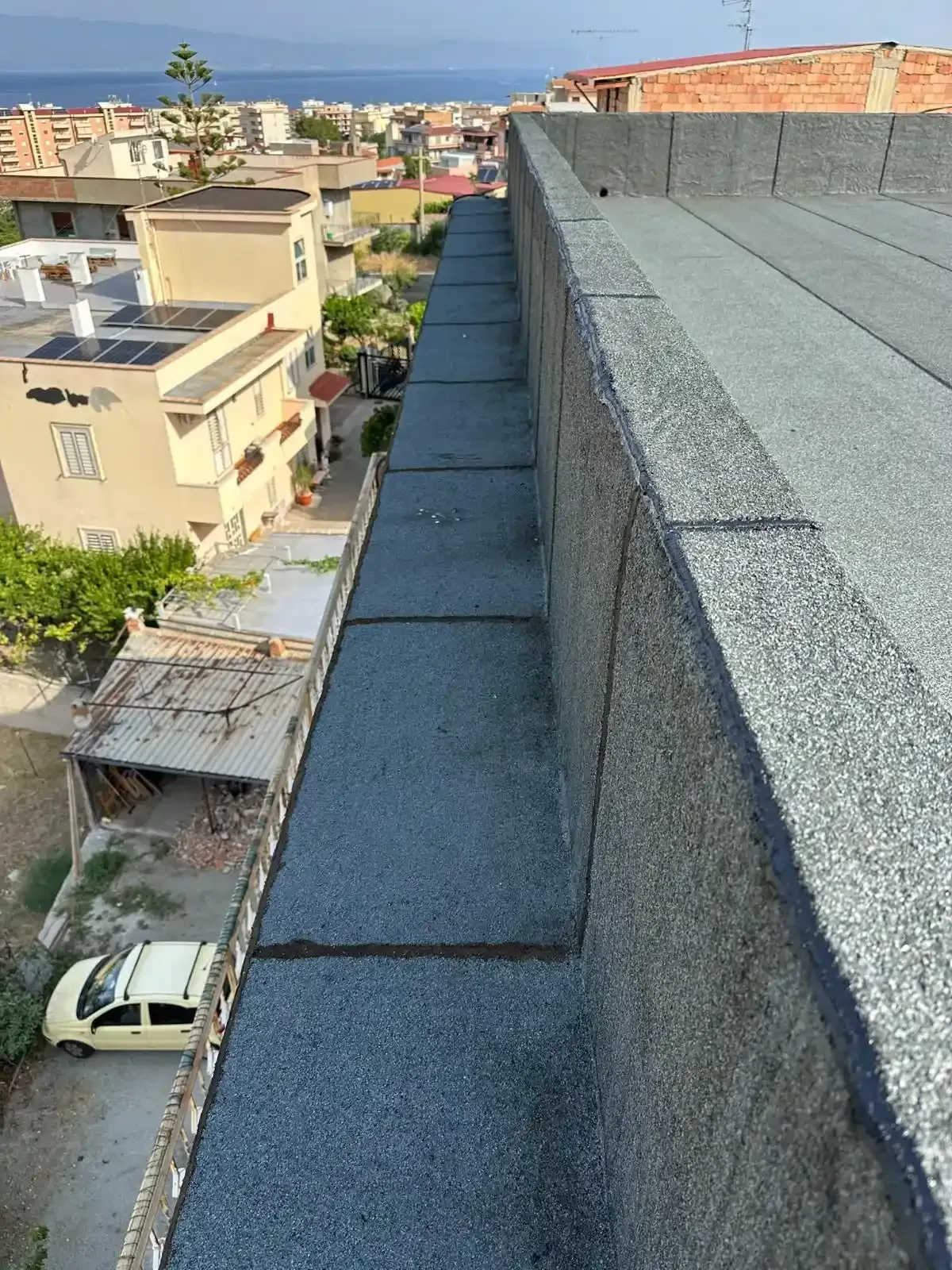 Vista di un tetto piano con tegole di asfalto grigio e un basso parapetto, con vista sulla città e sul mare.