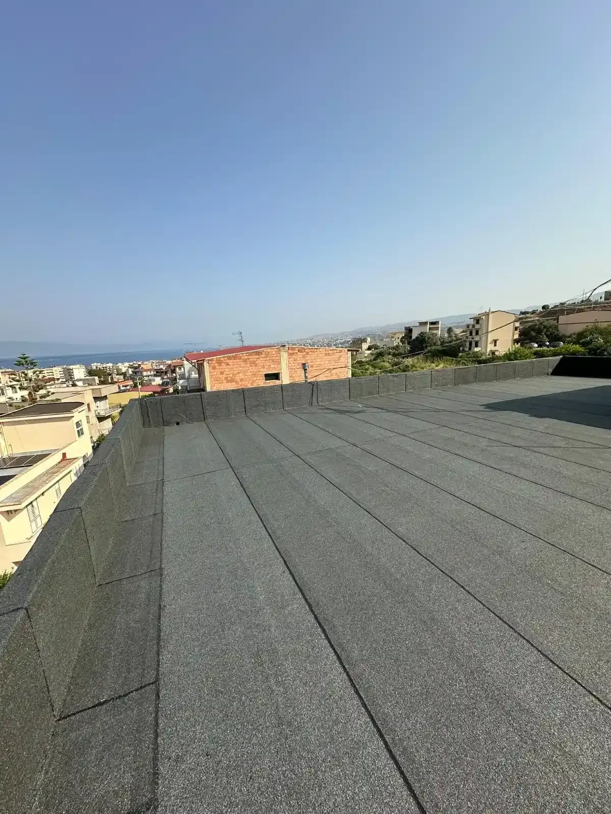 Tetto piatto e scuro con ghiaia grigia, con vista sulla città con mare e cielo azzurro.