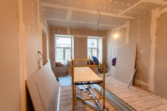 Stanza in fase di ristrutturazione; cartongesso installato su pareti e soffitto, impalcature al centro, due finestre sullo sfondo.