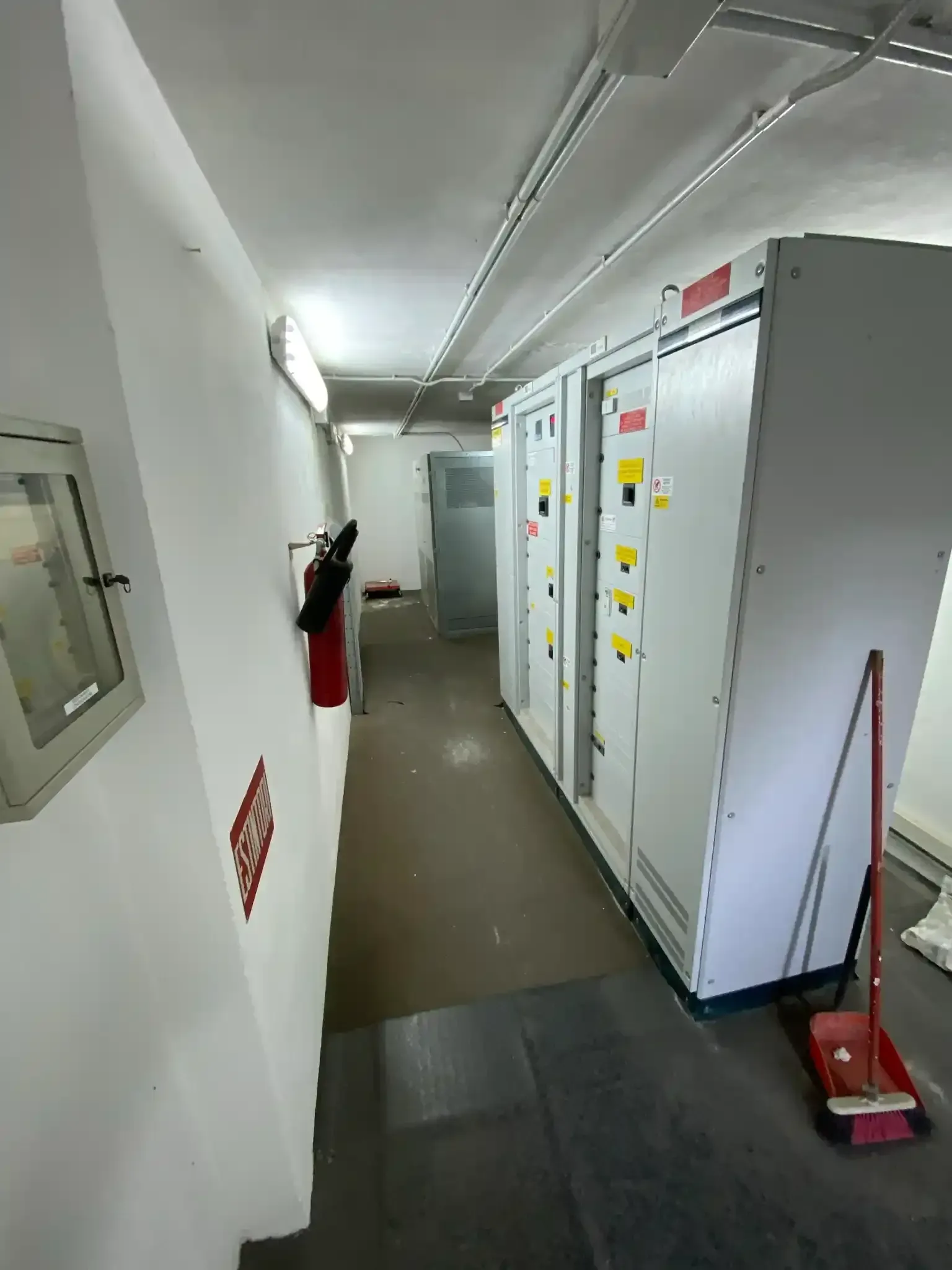 Locale elettrico con armadietti con pannelli grigi, estintore, scopa e corridoio stretto.