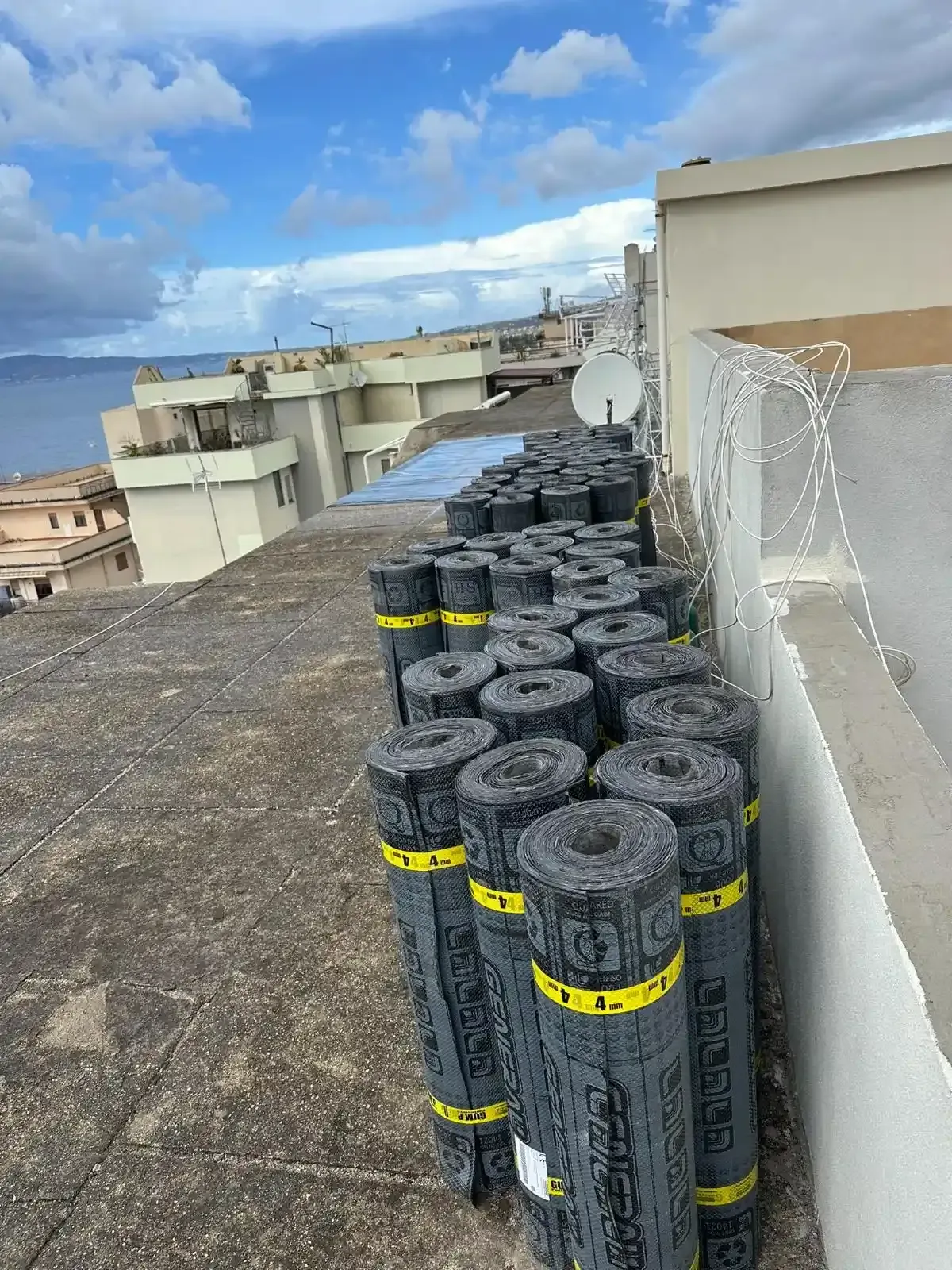 Rotoli di materiale di copertura su un tetto, con edifici e oceano visibili sullo sfondo.