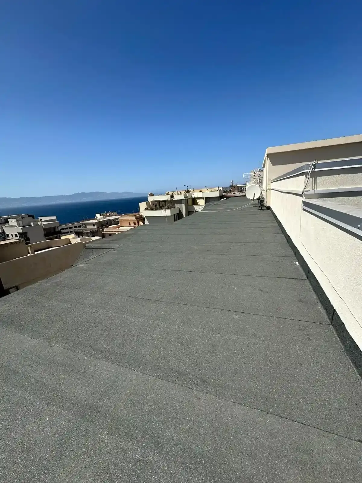 Tetto piatto, grigio scuro, con vista sugli edifici e sul mare sotto un cielo azzurro e terso.