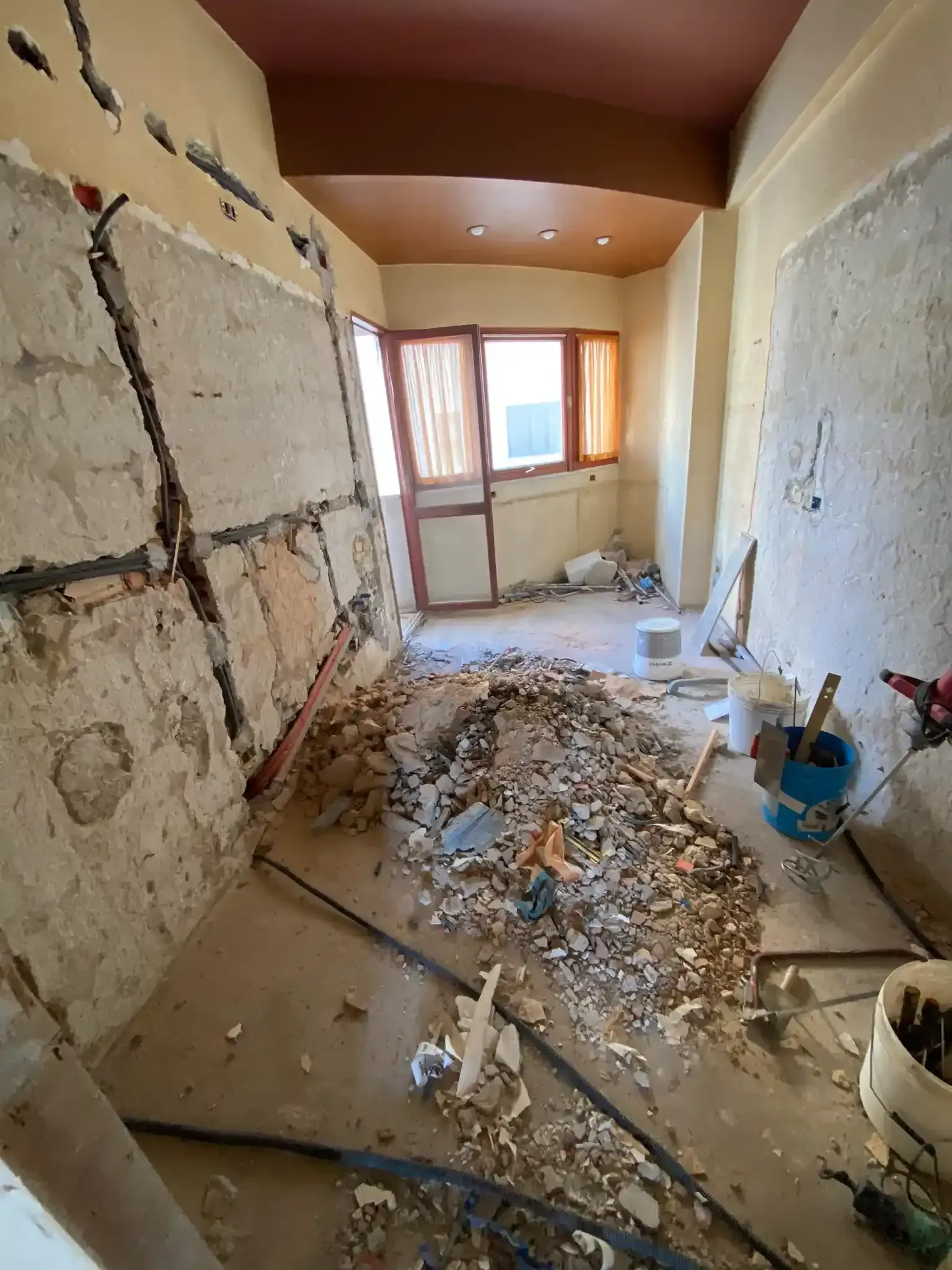 Stanza in fase di ristrutturazione, detriti sul pavimento, pareti parzialmente demolite, porta e finestra visibili.
