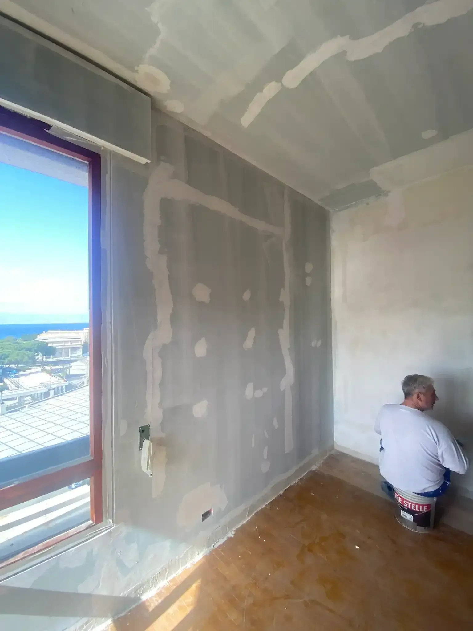 Un uomo applica il composto per cartongesso su una parete di una stanza. Finestra con vista sulla sinistra.