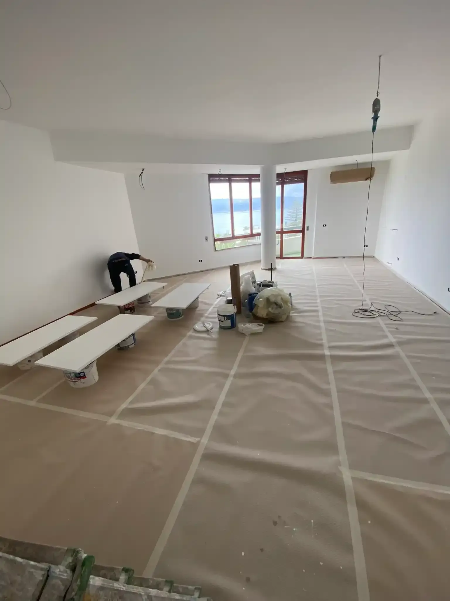 Una persona dipinge le finiture in una stanza in fase di ristrutturazione con vista sull'oceano. Pareti bianche, pavimento con rivestimento protettivo.