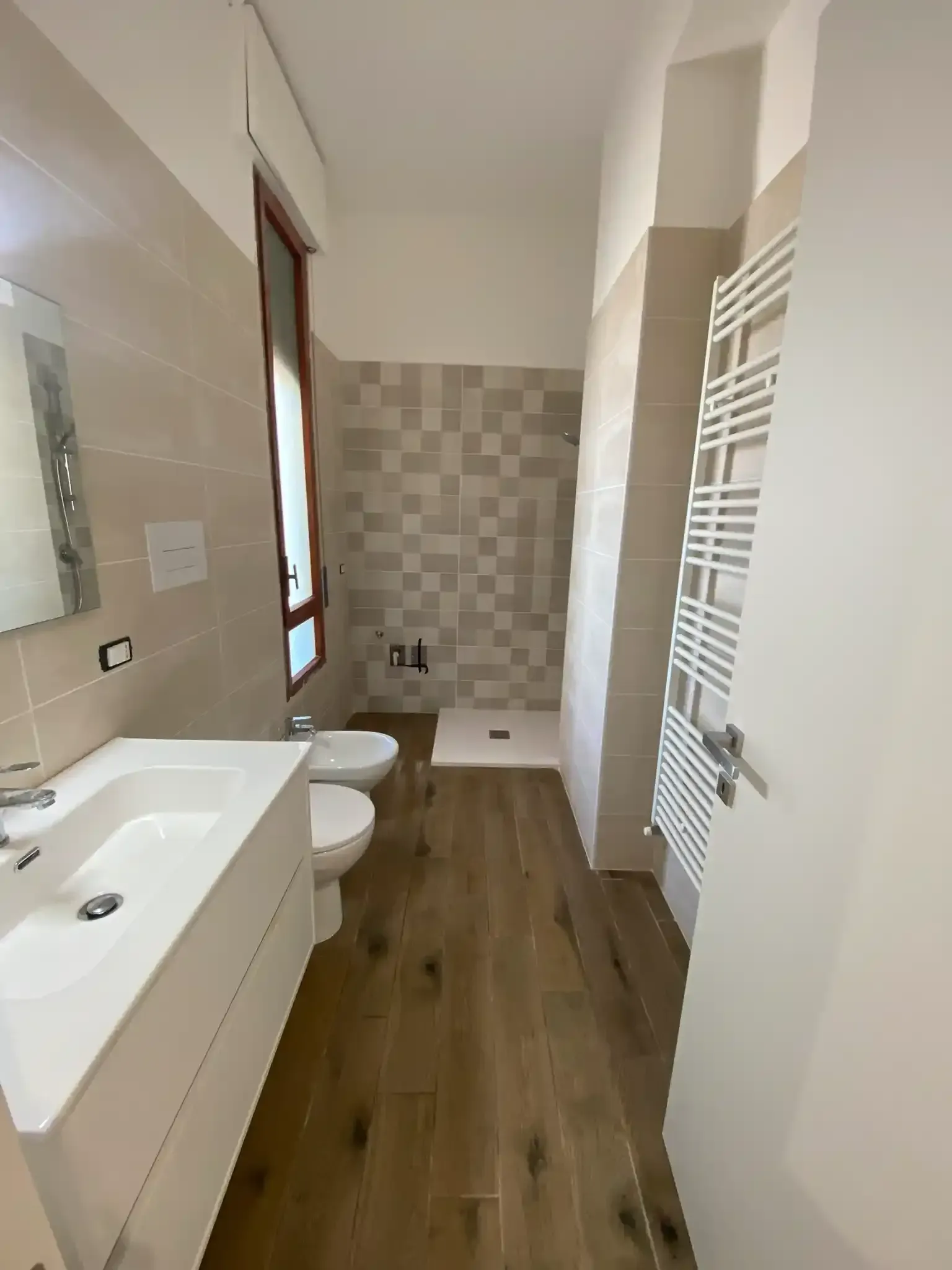 Bagno con lavandino, WC, bidet, doccia e scaldasalviette. Pavimento effetto legno, pareti dai toni neutri.