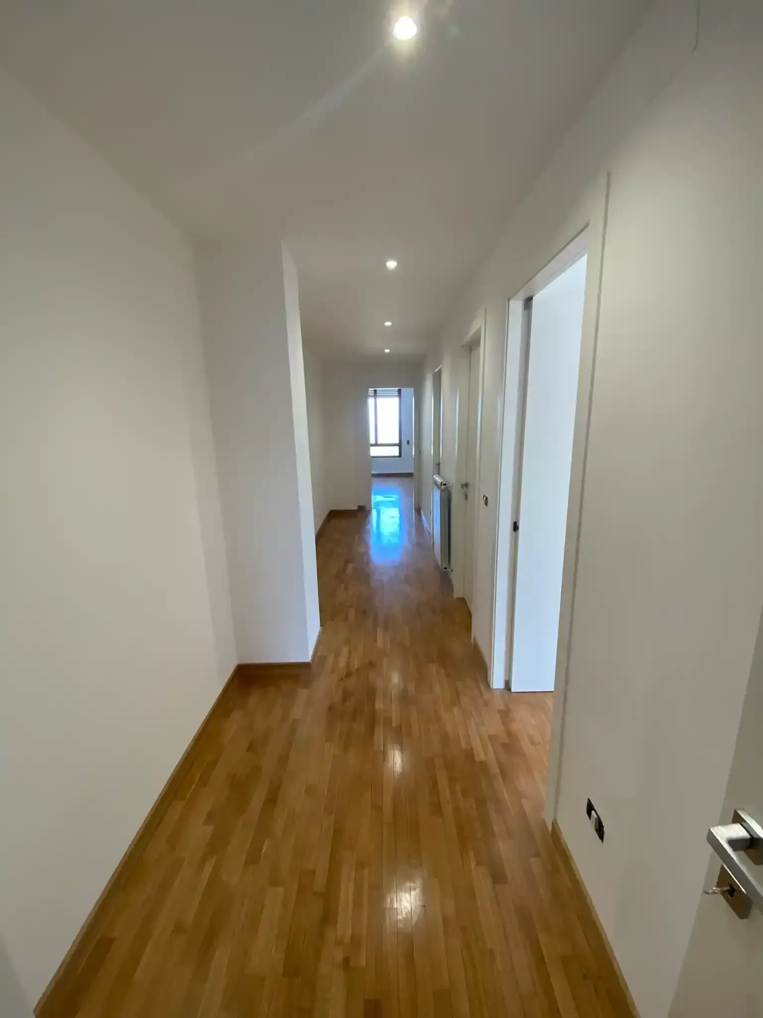 Lungo corridoio con pavimento in legno, pareti bianche e numerose porte chiuse.