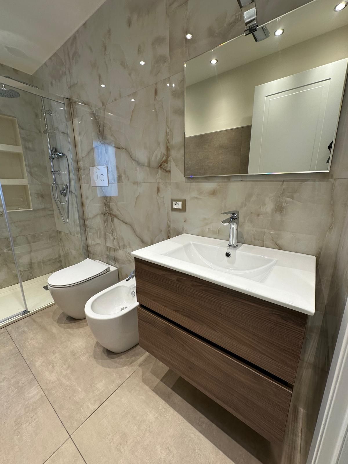 Bagno stretto con pareti piastrellate grigie, doccia, lavandini, WC, bidet e una porta in legno.