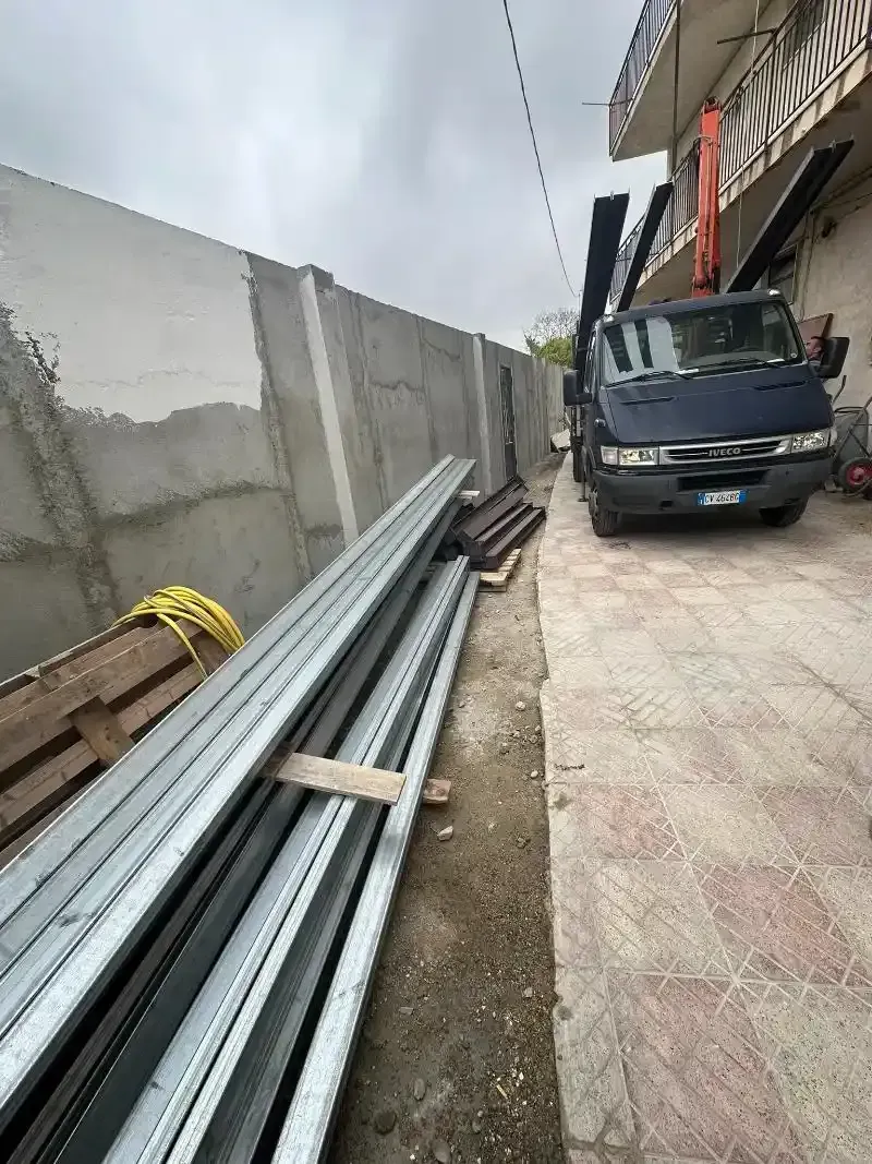 Travi metalliche accatastate accanto a un muro grigio e a un camion parcheggiato; materiali da costruzione su un'area pavimentata.