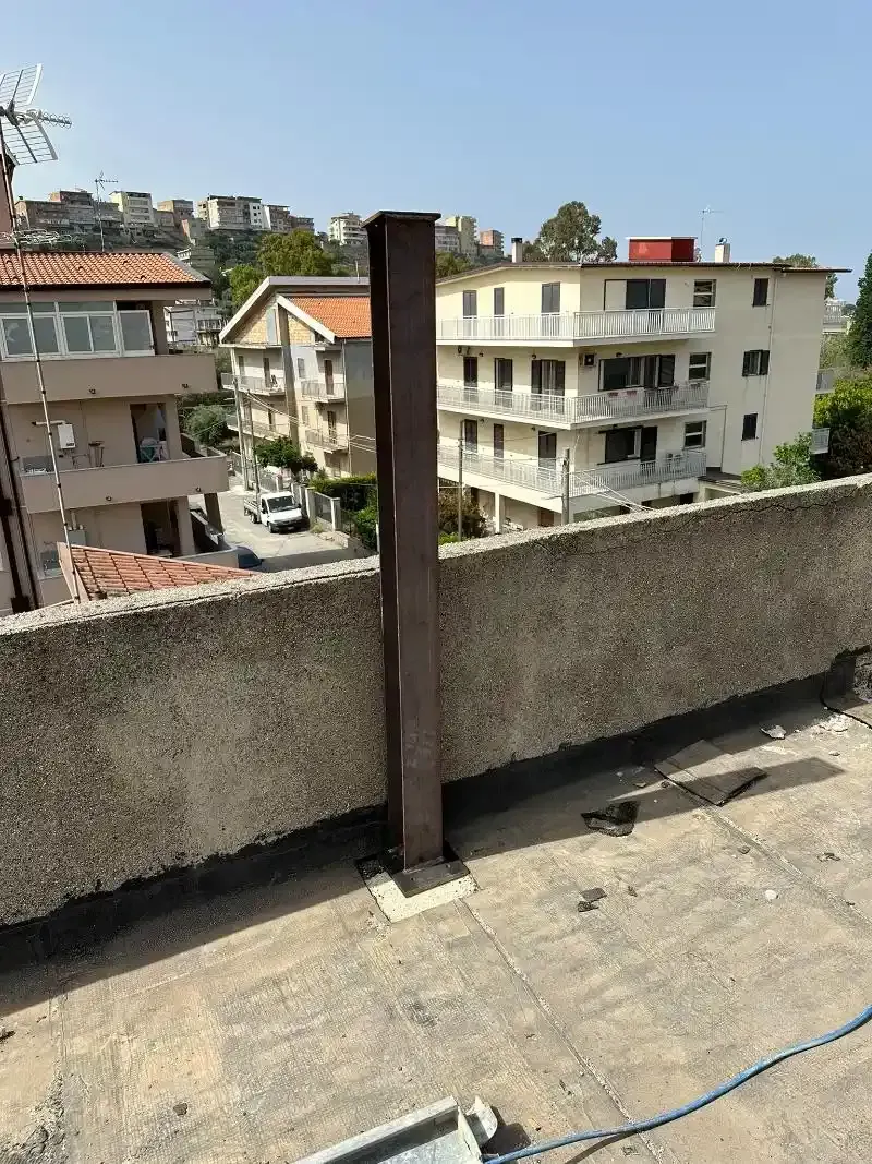 Un'alta trave di metallo marrone si erge su un tetto piatto con edifici sullo sfondo.