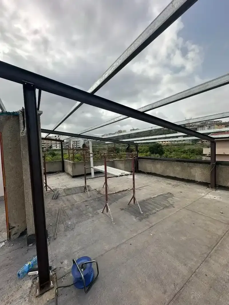 Costruzione di una tettoia con struttura metallica su un tetto. Il cielo è nuvoloso e il terreno è in cemento.