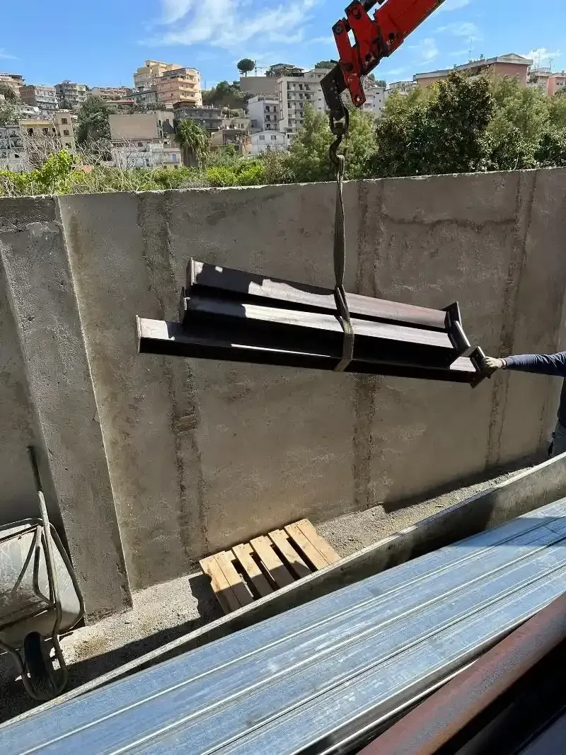 Una gru solleva una pila di travi di metallo scuro vicino a un muro di cemento; le travi di acciaio sono a terra.